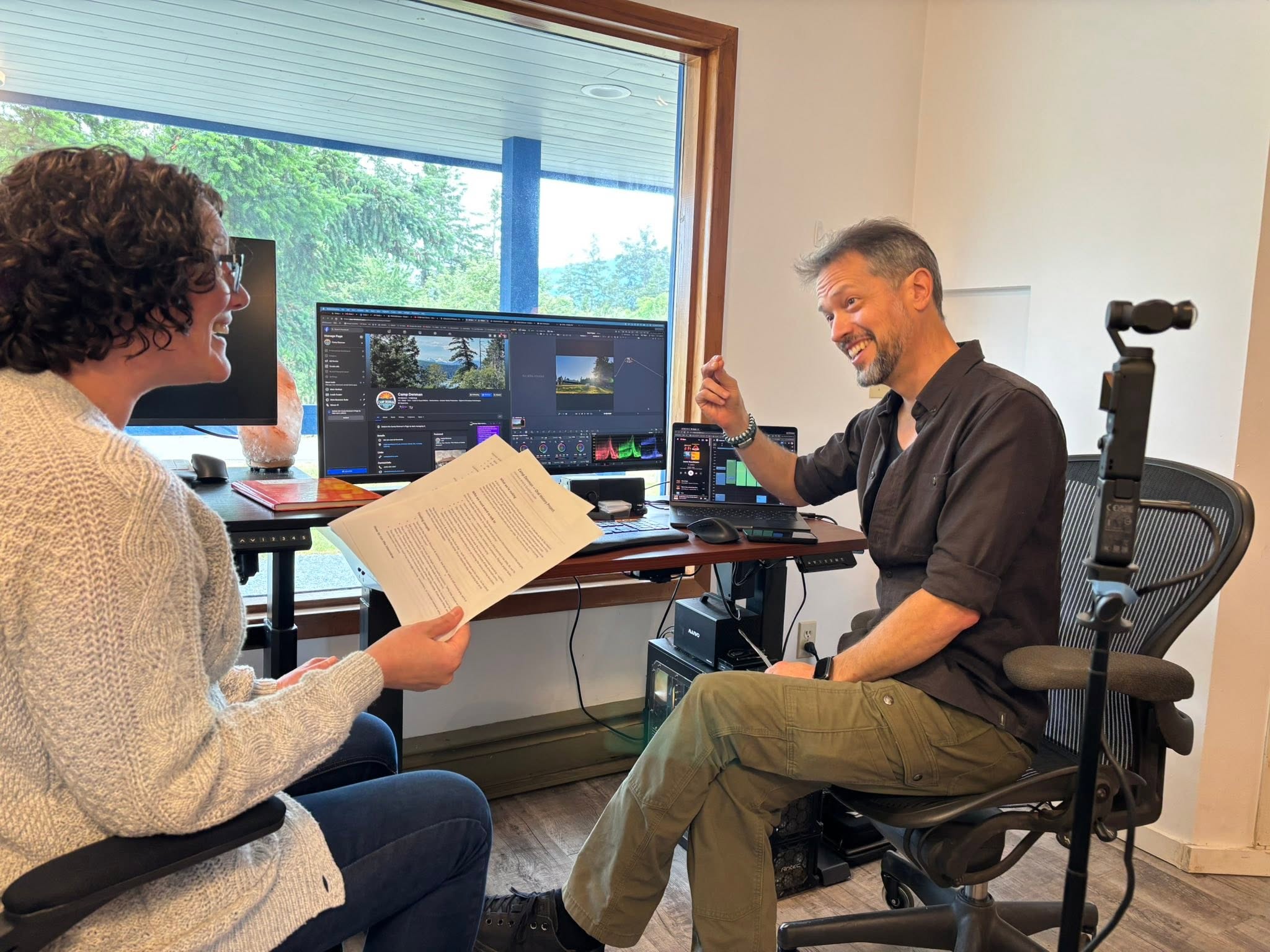 Two Camp Denman collaborators laughing over notes at an edit-suite desk overlooking the trees